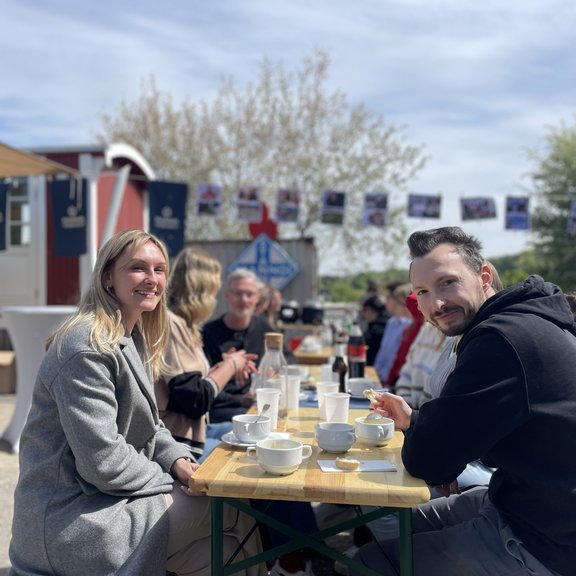 Personen sitzen an einem Tisch draußen und lachen in die Kamera.
