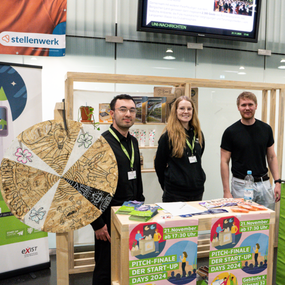 The picture shows two young men and a young woman in the centre. They are standing behind a small counter with a wheel of fortune to their left.