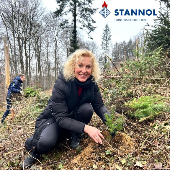 Ein blonde Frau pflanzt einen Baum im Wald. Sie trägt dunkle Kleidung und lächelt freundlich in die Kamera. Im Hintergrund ist eine weitere Person zu sehen, die einen Baum trägt.
