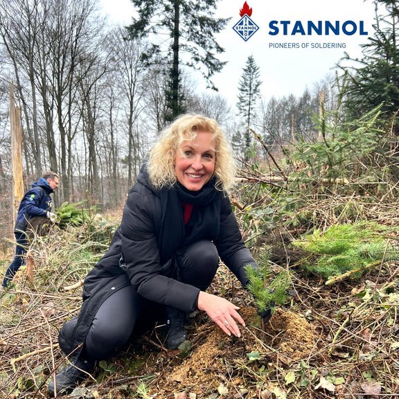 A blonde woman plants a tree in the forest. She is wearing dark clothes and smiling friendly into the camera. Another person carrying a tree can be seen in the background.