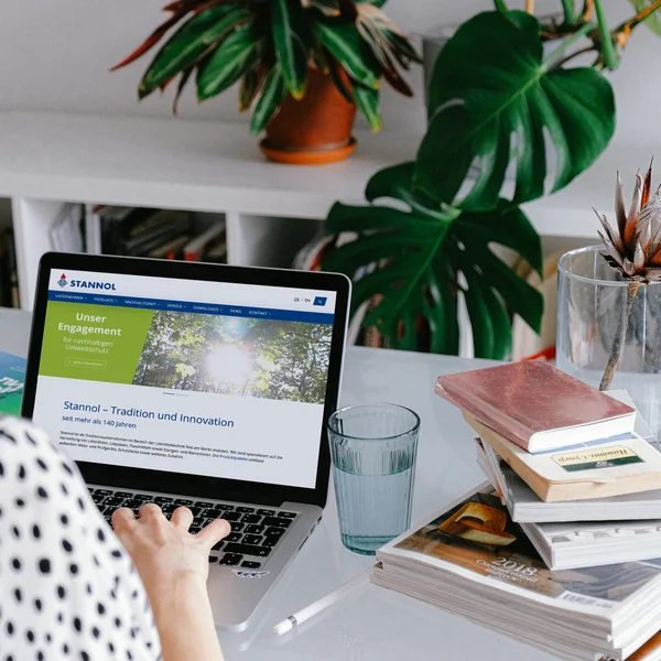 Auf dem Bild ist eine Frau im Anschnitt zu sehen, die vor einem Laptop sitzt. Daneben liegt ein Stapel Bücher auf dem Tisch. Im Hintergrund ist eine Pflanze zu sehen.