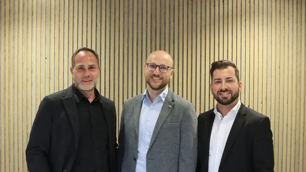 The picture shows three men in shirts and suits. They are smiling friendly into the camera.