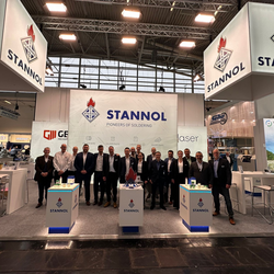 Auf dem Bild ist der Stannol-Messestand zu sehen. Im Vordergrund steht eine größere Personengruppe, die freundlich in die Kamera lächelt.