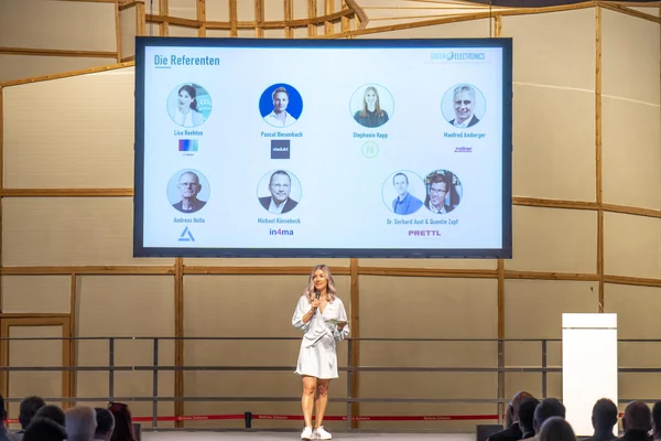 Auf dem Bild st die Moderatorin der Veranstaltung, Sabrina Nickel, auf der Bühne zu sehen. Sie hat blonde Haare und trägt ein knielanges Kleid. Auf dem Bildschirm über ihr sind die Porträts der Referenten und Referentinnen zu sehen.