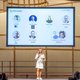 The picture shows the presenter of the event on stage: She is blonde and wears a knee-length dress. A screen with portraits of all the speakers can be seen in the background.