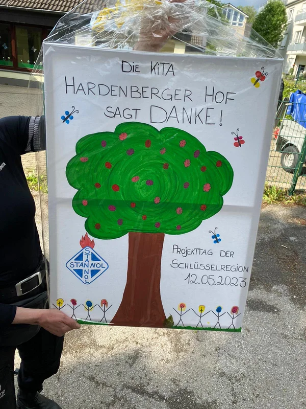 The image shows a person holding up a gift – a self-painted picture wrapped in clear plastic. The painting depicts a tree, the Stannol logo, butterflies, a group of children, and the text: "The Hardenberger Hof daycare says thank you! Project day Key Region 23.05.2023."