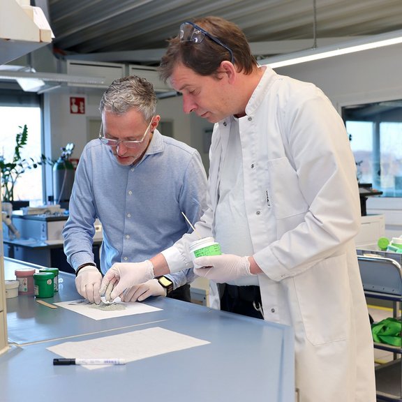 The picture shows two men in a laboratory experimenting with solder paste. The man on the left has grey hair, wears glasses and a light blue shirt. The man on the right has brown hair and is wearing a white coat.