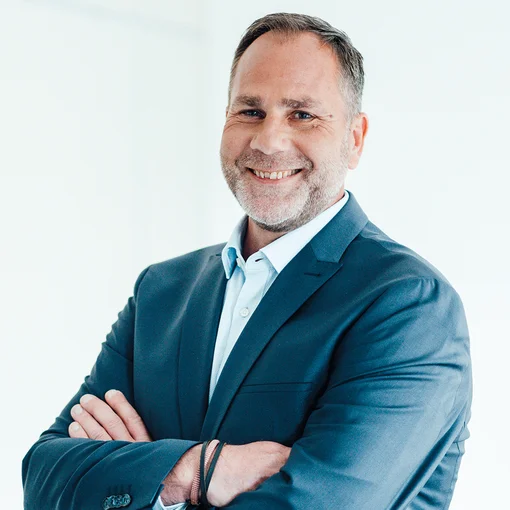 Auf dem Foto ist ein Mann in einem hellen Hemd und einem blauen Sakko zu sehen. Er hat leicht grauen Haare und einen leicht grauen Bart. Er lächelt freundlich in die Kamera.