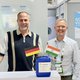 Two men are smiling at a trade show booth with German and Indian flags.