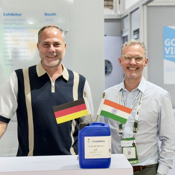 Auf dem Bild sind zwei Männer zu sehen, die freundlich in die Kamera lächeln. Im Vordergrund steht ein blauer Stannol-Flussmittelkanister mit der indischen und deutschen Flagge.