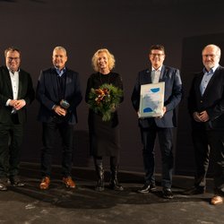 Auf dem Bild sind fünf Personen vor einem dunklen Hintergrund zu sehen. Drei von ihnen halten eine Urkunde eine Trophäe und einen Blumenstrauß in den Händen.