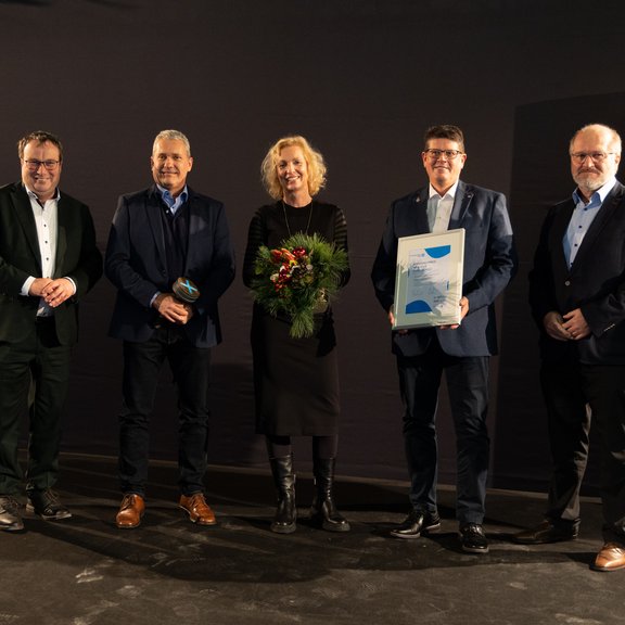 Auf dem Bild sind fünf Personen vor einem dunklen Hintergrund zu sehen. Drei von ihnen halten eine Urkunde eine Trophäe und einen Blumenstrauß in den Händen.