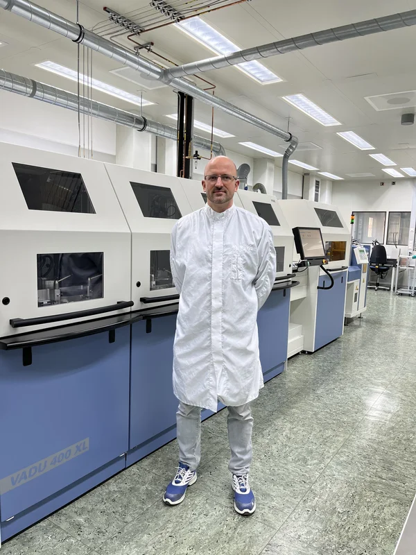 Mann in Schutzanzug steht in einem Labor mit Maschinen im Hintergrund.