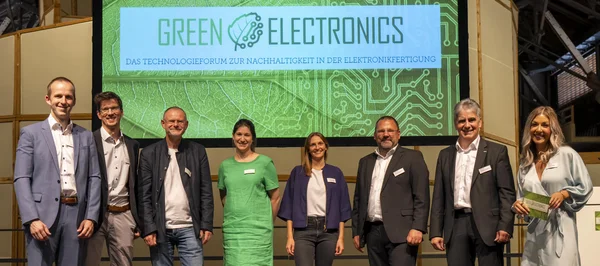 The picture shows eight people, five men and three women. They are standing in front of a large screen on which the logo of the ‘Green Electronics’ event has been displayed. They are all smiling friendly into the camera.