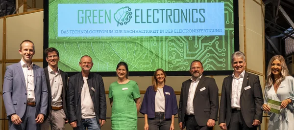 Auf dem Bild sind acht Personen zu sehen, fünf Männer und drei Frauen. Sie stehen vor einem großen Bildschirm, auf dem das Logo der Veranstaltung "Green Electronics" eingeblendet wurde.
