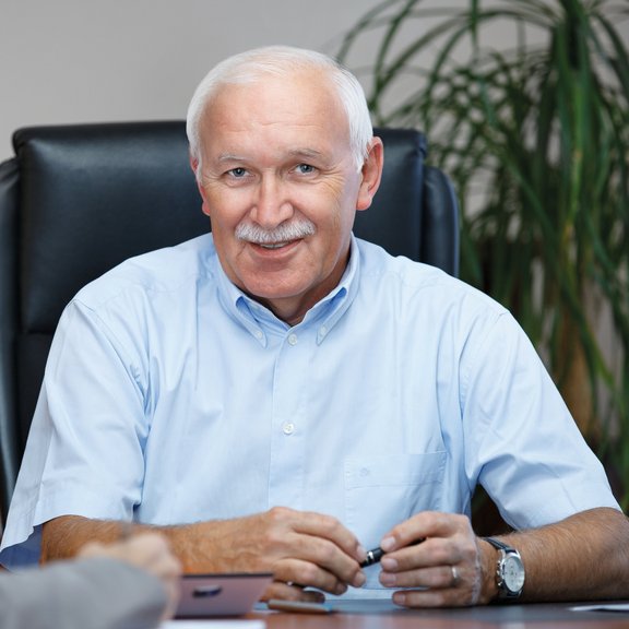 The picture shows former Managing Director Karl-Heinz Dörr. He has white hair, a moustache and is wearing a light blue shirt. He is sitting at a desk and holding a pen in his hand.