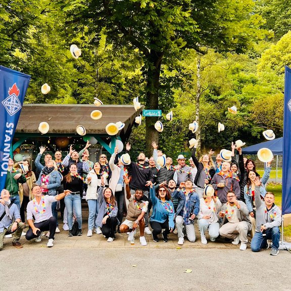 The picture shows a large group of people throwing straw hats into the air. In the background you can see trees and a barbecue hut.