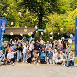 Auf dem Bild ist eine größere Gruppe von Menschen zu sehen, die Strohhüte in die Luft werfen. Im Hintergrund sieht man Bäume und eine Grillhütte.