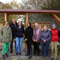 Auf dem Bild ist eine Gruppe von sieben Männern und Frauen vor einem überdachten Insektenhotel zu sehen.