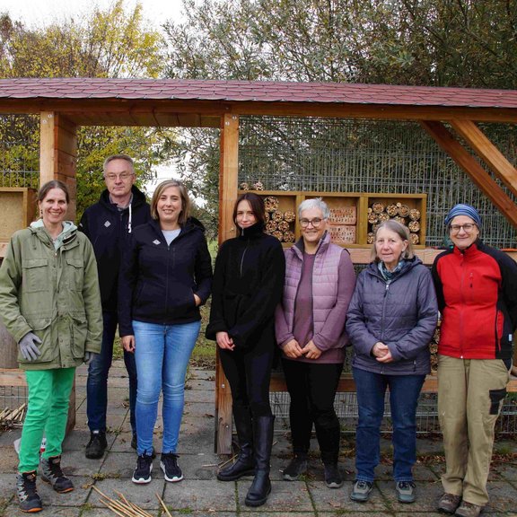 Auf dem Bild ist eine Gruppe von sieben Männern und Frauen vor einem überdachten Insektenhotel zu sehen.