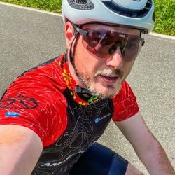 Auf dem Bild ist ein junger Mann mit weißem Fahrradhelm, rot-schwarzem Trikot und Radbrille auf einem Rennrad zu sehen. Er macht ein Selfie und lächelt freundlich in die Kamera.