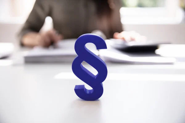 The picture shows a blue plastic paragraph sign. It is standing on a table, with a person sitting at the table in the background.