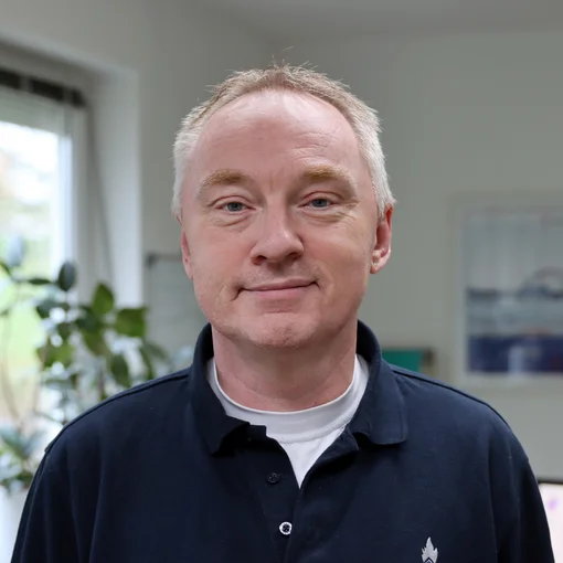 The picture shows a man with thin, grey hair. He is wearing a dark blue shirt with a white T-shirt underneath. He smiles friendly at the camera.