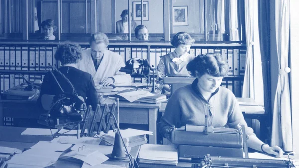 he image shows the old office spaces of Stannol, presumably from the 1950s. It is presented in a nostalgic blue tone, enhancing the historical atmosphere. In the foreground, a woman is seated at a desk, working intently on documents. Additional employees can be seen in the background, also engaged in office tasks. The furnishings consist of simple wooden furniture, including desks and shelves filled with files and folders. Large windows provide natural light to the room. The scene offers an impression of the work environment and daily life in an office during that era.