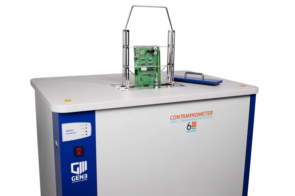 Auf dem Bild ist ein Contaminometer von GEN3 Systems zu sehen, ein Prüfgerät für Baugruppen auf leitfähige Rückstände. Zu sehen ist eine graue, kastenförmige Apparatur mit einem Halter für Leiterplatten. Die Leiterplatten können in das Gerät abgesenkt und dort überprüft werden. 