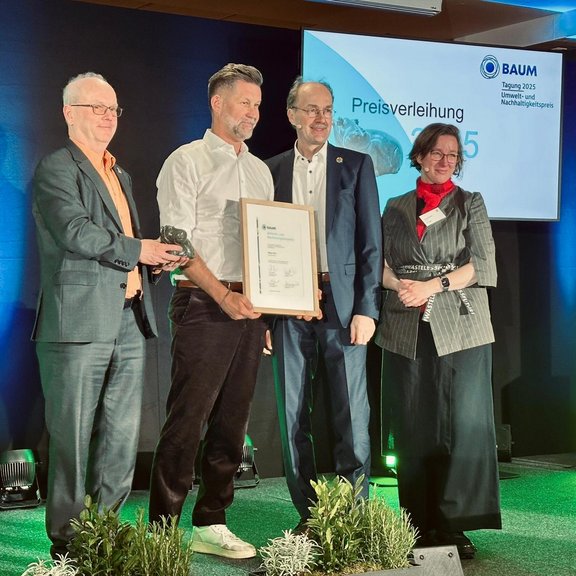 Vier Personen auf einer Bühne, eine Person hält eine Urkunde.