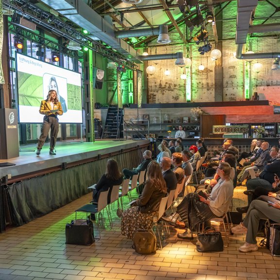 Auf dem Bild ist eine Bühne und ein Zuschauerraum in einer alten Fabrikhallezu sehen. Auf der Bühne steht eine junge blonde Frau, vor der Bühne sind mehrere belegte Stuhlreihen zu sehen.