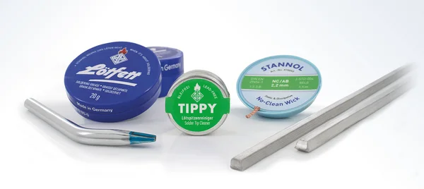 Auf dem Bild ist verschiedenes Zubehör zu sehen, wie eine runde, blaue Dose Lötfett, eine kleine, runde metallene Dose mit Tippy-Lötspitzenreiniger, eine Ersatz-Lötspitze und eine Rolle mit Entlötlitze.