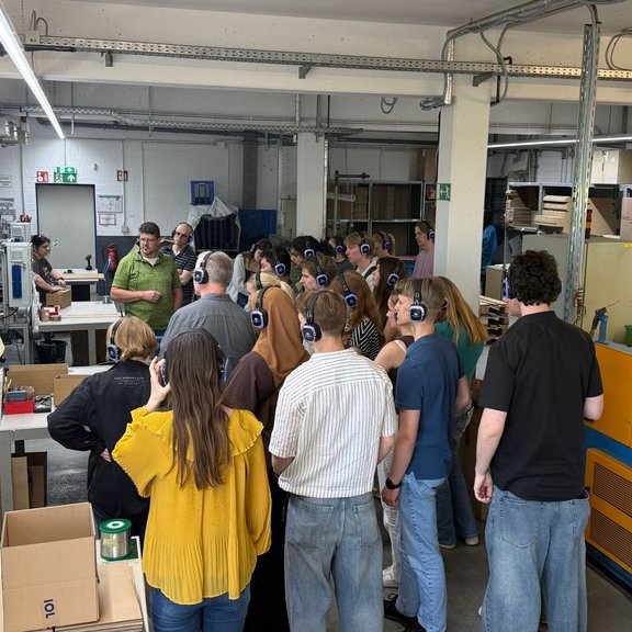 Personengruppe mit Kopfhörern in einem industriellen Raum, hört einen Vortrag.