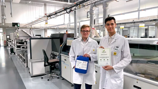 Auf dem Bild sind zwei junge Männer in einer Produktionshalle zu sehen. Sie tragen beide weiße Kittel. Der Mann links ist blond und hält einen blauen Flussmittel-Kanister in den Händen. Der Mann rechts hat dunkle Haare und hält ein Zertifikat in die Kamera.