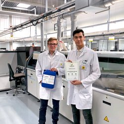 Auf dem Bild sind zwei junge Männer in einer Produktionshalle zu sehen. Sie tragen beide weiße Kittel. Der Mann links ist blond und hält einen blauen Flussmittel-Kanister in den Händen. Der Mann rechts hat dunkle Haare und hält ein Zertifikat in die Kamera.