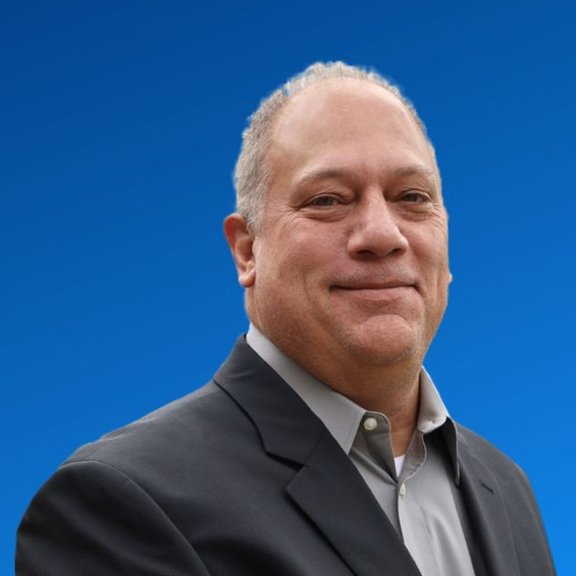 The picture shows a middle-aged man in a shirt and jacket against a blue background. He smiles friendly into the camera.