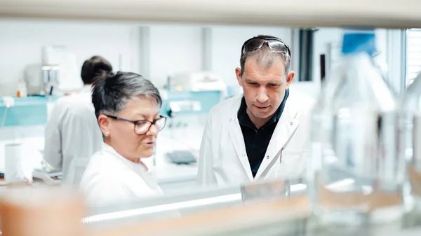 Auf dem Foto ist eine Labormitarbeiterin und ein Labormitarbeiter im Gespräch zu sehen. Sie tragen beide weiße Kittel.
