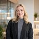 Blonde Frau in Büro mit schwarzem Blazer, lächelt in die Kamera.