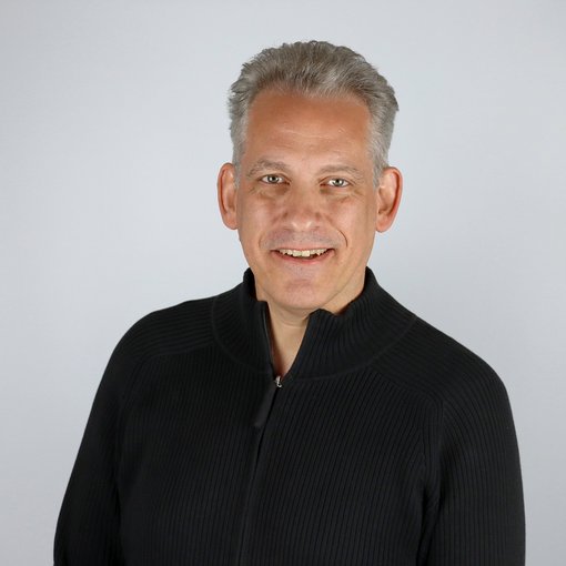 Auf dem Bild ist ein Mann in einem dunklen Pullover zu sehen. Er hat graue Haare und lächelt freundlich in die Kamera.