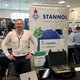 The picture shows a young man in a white shirt standing behind a small exhibition stand. To his right is a Stannol display with various products. 