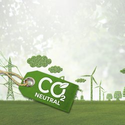 Das Bild zeigt eine symbolische Darstellung von Nachhaltigkeit und Klimaneutralität. Im Vordergrund befindet sich ein grünes Etikett mit der Aufschrift „CO₂ Neutral“. Das Etikett ist mit einer Schnur befestigt und hat ein Blatt-Symbol im Schriftzug, was auf umweltfreundliche und klimafreundliche Aspekte hinweist. Im Hintergrund erstreckt sich eine grüne Landschaft mit verschiedenen Elementen, die aus Pflanzen und Bäumen geformt sind. Dazu gehören Windkraftanlagen, Strommasten und Gebäude, die alle aus Laub oder Gras gestaltet sind. Es sind außerdem Wolken aus Blattstrukturen zu sehen. Der Hintergrund ist leicht verschwommen und vermittelt eine friedliche, naturverbundene Atmosphäre. Das Bild symbolisiert den Fokus auf erneuerbare Energien und CO₂-Neutralität.