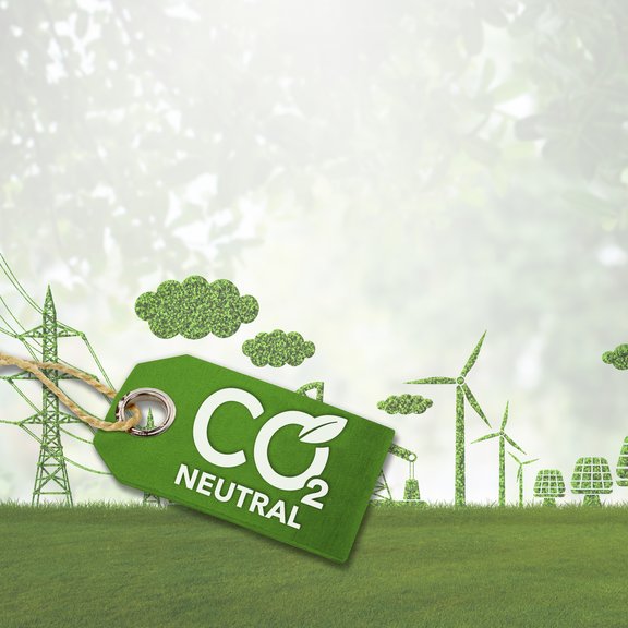 The image shows a symbolic representation of sustainability and climate neutrality. In the foreground, there is a green tag with the inscription "CO₂ Neutral." The tag is attached with a string and includes a leaf symbol within the lettering, emphasizing eco-friendly and climate-friendly aspects. In the background, a green landscape stretches out with various elements made of plants and trees. These include wind turbines, power poles, and buildings, all crafted from foliage or grass. Additionally, there are clouds made of leafy structures. The background is slightly blurred, creating a peaceful, nature-oriented atmosphere. The image symbolizes a focus on renewable energy and CO₂ neutrality.