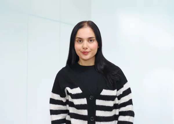 Auf dem Bild ist ein junge Frau mit dunklen, glatten Haaren, einem schwarzen Top und einer weiß-schwarz-gestreiften Strickjacke zu sehen. 