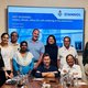 The picture shows a group of people in front of a screen with a PowerPoint presentation on Stannol. Marco Dörr, Thomas Kolossa and Batcheva Dominguez-Tellez are standing or sitting in the centre. They are surrounded by their Indian hosts. All of them are smiling friendly into the camera. 