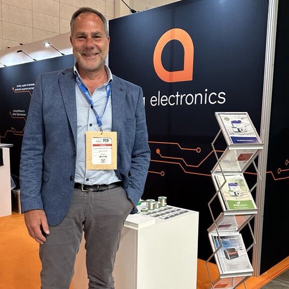 The picture shows a middle-aged man standing in front of a trade fair stand and smiling friendly into the camera.