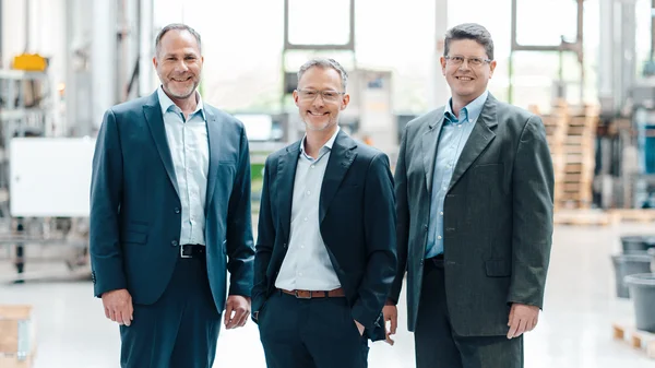 The image shows the three authorized signatories of Stannol (from left to right): Thomas Kolossa, Ingo Lomp, and Herbert Schmidt. All three are wearing formal attire: Thomas Kolossa and Ingo Lomp are wearing dark blue blazers with light blue shirts, while Herbert Schmidt is wearing a charcoal-colored blazer with a light blue shirt. They are standing next to each other and looking friendly at the camera. In the blurred background, the production environment of Stannol can be seen, giving the image a professional context. Overall, the image conveys a professional yet friendly impression.