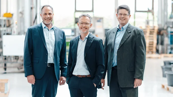 Das Bild zeigt die drei Prokuristen von Stannol (vlnr): Thomas Kolossa, Ingo Lomp und Herbert Schmidt. Alle drei tragen formelle Kleidung: Thomas Kolossa und Ingo Lomp tragen ein dunkelblaues Sakko mit einem hellblauen Hemd und Herbert Schmidt ein anthrazitfarbenes Sakko mit einem hellblauen Hemd. Sie stehen nebeneinander und blicken freundlich in die Kamera. Im unscharfen Hintergrund ist die Produktionsumgebung von Stannol zu sehen, was dem Bild einen professionellen Rahmen verleiht. Insgesamt vermittelt das Bild einen professionellen und zugleich freundlichen Eindruck.