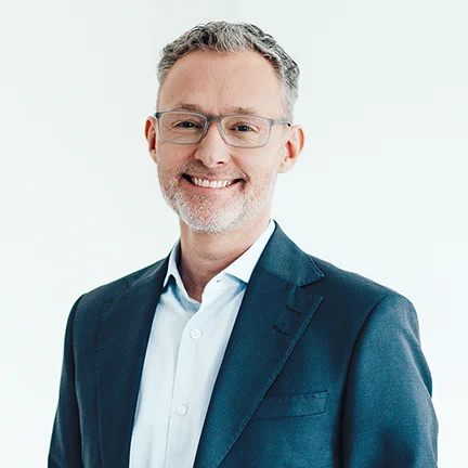 Auf dem Bild ist ein Mann mit hellem Hemd und blauem Sakko zu sehen. Er hat kurze graue Haare und trägt eine Brille. Er lächelt freundlich in die Kamera.