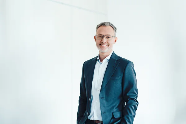 Auf dem Bild ist ein Mann in hellem Hemd und blauem Sakko zu sehen. Er hat kurze graue Haare, trägt eine Brille und lächelt freundlich in die Kamera.