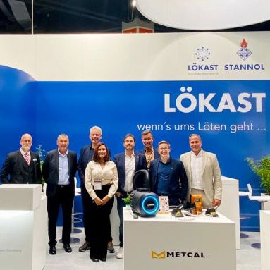 The picture shows a group of people in formal attire at a trade fair stand, smiling friendly into the camera. The Stannol and Lökast logos can be seen in the background.